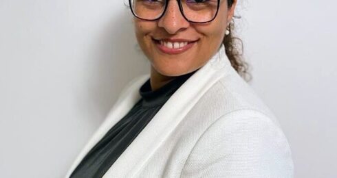 Adan Azem stands against a white background, wearing a white blazer and black top. She is wearing black-rimmed glasses.