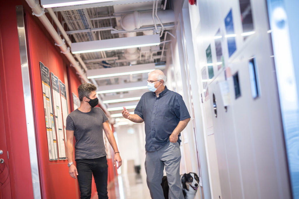 Andrea and Pinder walk down the red hallway on the third floor of QMI with Hunter the dog
